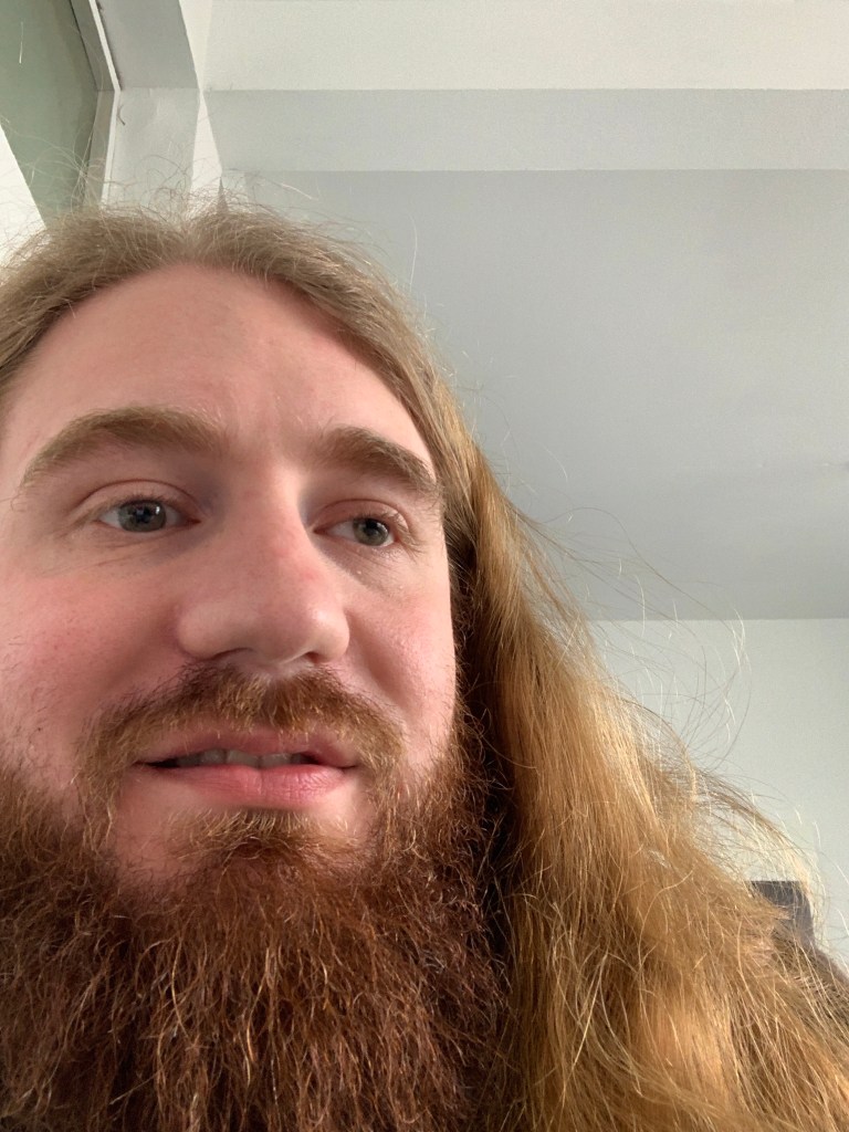 Ich habe auf diesem Bild lange Haare, einen Bart und lächel leicht in die Kamera ￼￼
