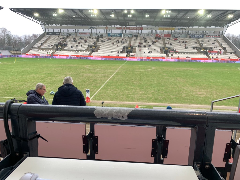 Ich sitze im Stadion an der Hafenstraße und habe dabei ein Foto geschossen von einer Tribüne, wo davor der Fußballplatz zu sehen ist, ￼￼￼