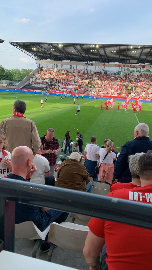 Das Bild zeigt eine lebhafte Szene in einem Fußballstadion während eines Spiels. Es ist taghell und das Wetter scheint klar und sonnig zu sein, denn der Himmel ist blau und hinter den Scheinwerfern der Stadionbeleuchtung sind keine Wolken zu erkennen. Der Blick ist von einer Tribüne hinter dem Tor aus aufgenommen, und wir sehen den hinteren Teil des Spielfelds, wobei der Großteil der Spielfläche von Zuschauern im Vordergrund verdeckt wird.

Die Tribünen sind gut gefüllt mit Zuschauern, die überwiegend rot-weiße Farben tragen, was darauf hindeutet, dass wir uns in einem Stadion befinden, dessen Heimteam diese Farben hat. Links im Bild ist eine größere, überdachte Tribüne zu sehen, die auch fast durchgehend besetzt ist. Auf der Tribüne sind zahlreiche Zuschauer in unterschiedlichen Posen zu sehen: Einige sitzen, andere stehen, und viele sind in Gespräche mit ihren Nachbarn vertieft oder konzentriert auf das Geschehen auf dem Spielfeld.

Im Vordergrund, zwischen den oberen und unteren Tribünenrängen, bewegen sich einige Personen. Ein Offizieller in schwarzer Kleidung beobachtet das Spiel aufmerksam und hält eine Fahne in der Hand, was auf einen Linienrichter hindeutet. Auf dem Spielfeld tragen die Spieler überwiegend weiße und rote Trikots, was auf zwei Mannschaften schließen lässt, eine in Weiß und die andere in Rot. Der Ball ist im Moment nicht sichtbar, aber die Spieler scheinen sich gerade auf eine Spielsituation vorzubereiten oder auf sie zu reagieren.

Was die Atmosphäre im Stadion betrifft, so wirkt sie begeistert und engagiert, wie es typisch für ein Fußballspiel mit leidenschaftlichen Anhängern ist. Das Grün des Rasens hebt sich deutlich von den hellen Farben der Fans und den dunklen Sitzreihen der Tribüne ab. Auf einem Schild an der überdachten Tribüne steht "STADION AN DER HAFENSTRASSE", was darauf hinweist, dass das Stadion möglicherweise diesen Namen trägt oder sich in der Nähe der Hafenstraße befindet. An der Wand der überdachten Tribüne sind zudem Werbeplakate und Logos zu sehen, was typisch ist für moderne Sportstätten, die Sponsoren präsentieren.