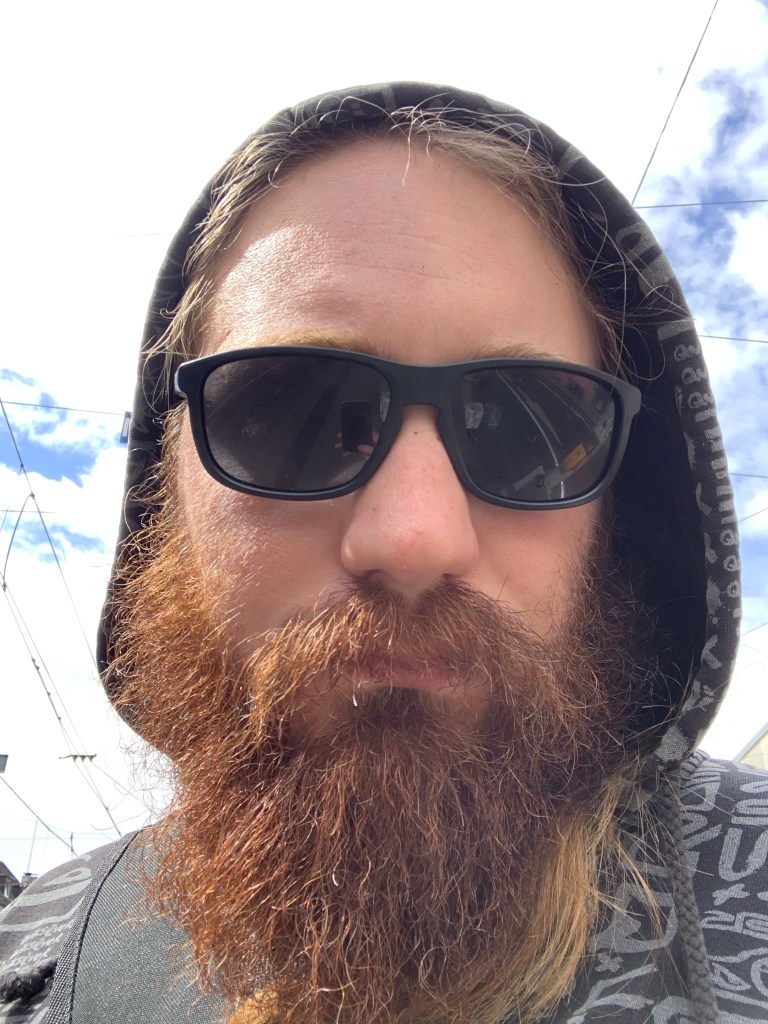 Auf dem Bild ist Jan zu sehen, der eine Sonnenbrille und eine Kapuze trägt. Hinter ihm kann man aus dem Fenster schauen und man sieht den Himmel.