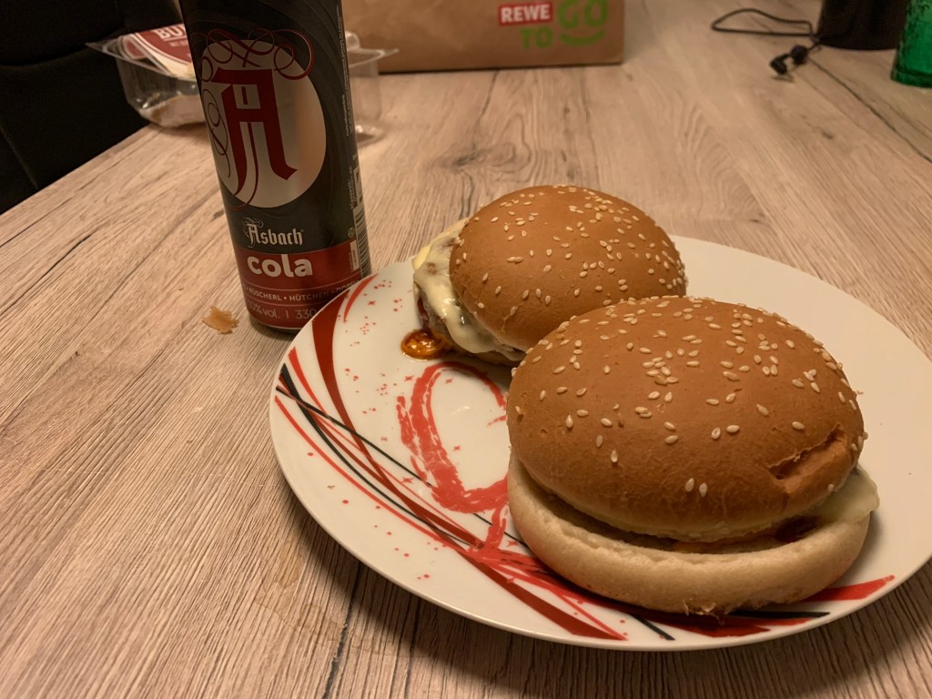 Auf einem Holztisch steht ein Teller mit zwei Burger darauf daneben eine Dose Asbach Cola und im Hintergrund eine ReWE Einkaufstüte